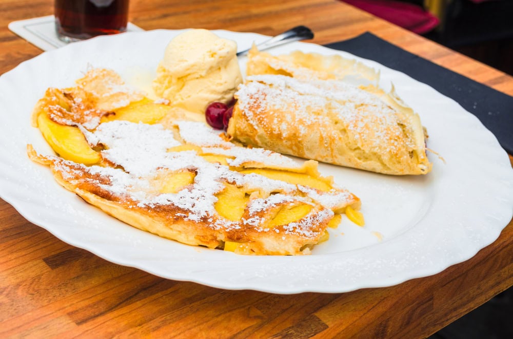 A white plate with apple strudel, a scoop of vanilla ice cream, a cherry, and a slice of pastry, all dusted with powdered sugar, on a wooden table.