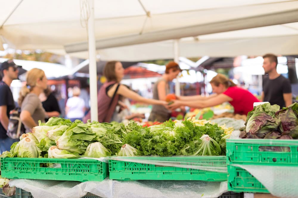 Fresh leafy vegetables in green crates at an outdoor market stand, with people shopping and interacting under white tents in the background.
