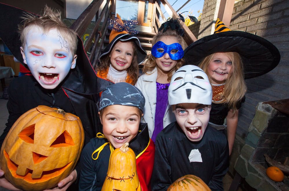 Six children in various Halloween costumes, including a vampire, witch, and masked characters, smiling and holding carved pumpkins indoors.