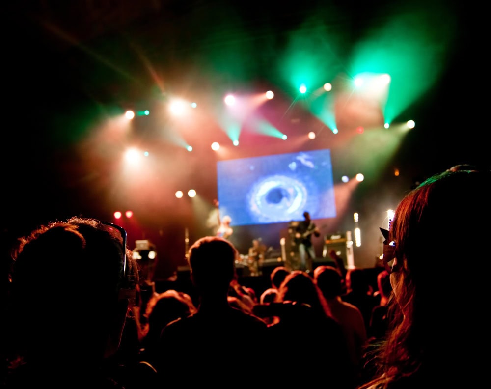 Audience watches a live concert with colorful stage lights and a large screen displaying a circular graphic behind the performers.