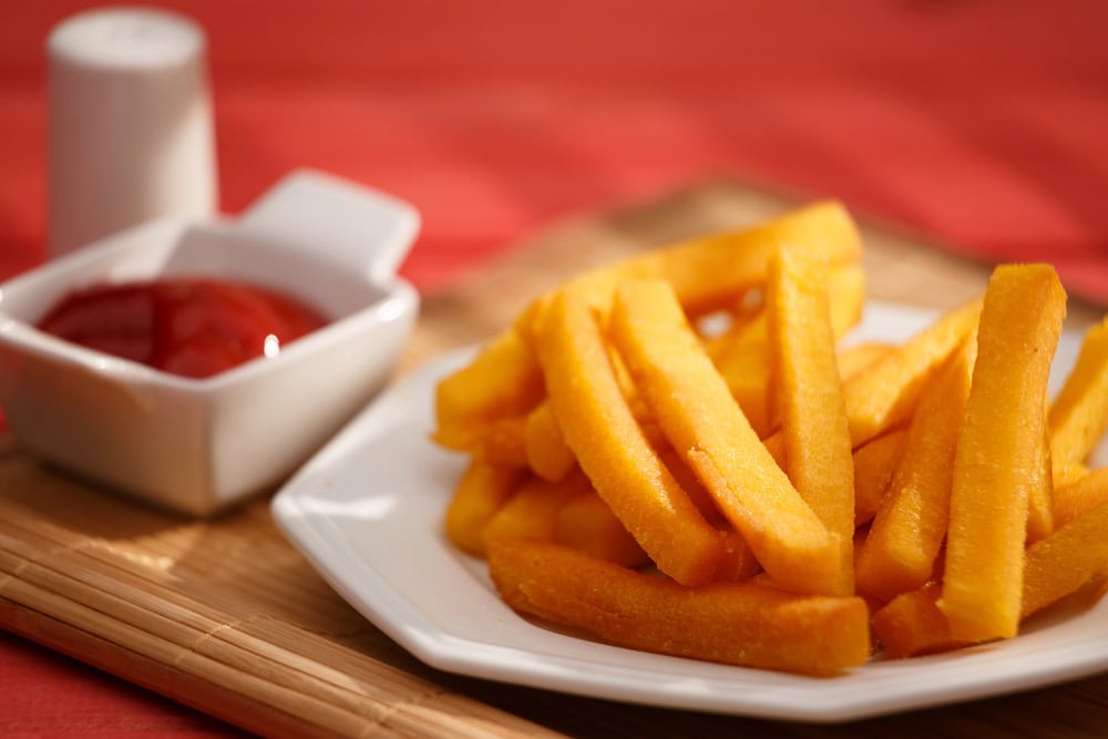 A white plate with French fries next to a small dish of ketchup and a salt shaker on a wooden mat with a red background.