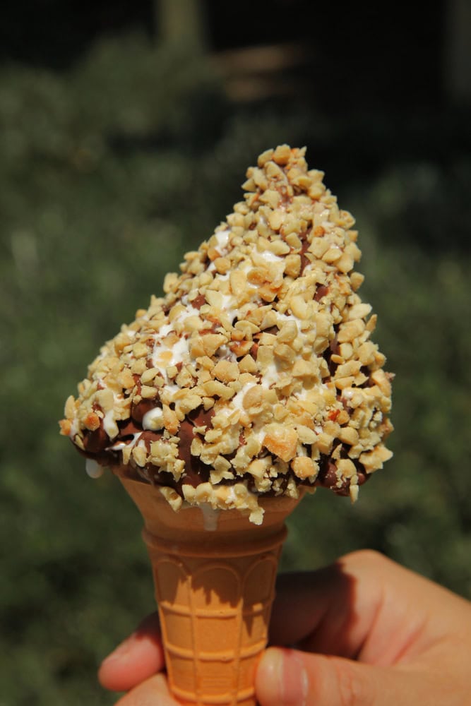 A hand holding an ice cream cone coated with chopped nuts and chocolate against a blurred outdoor background.