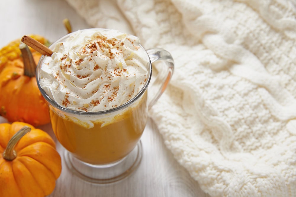 A glass mug of pumpkin spice latte topped with whipped cream and a cinnamon stick sits on a white table next to small pumpkins and a knitted cream-colored blanket.