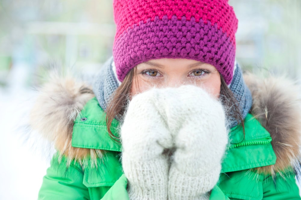 A person in a green coat and red knit hat holds gloved hands to their face outdoors in a snowy setting.