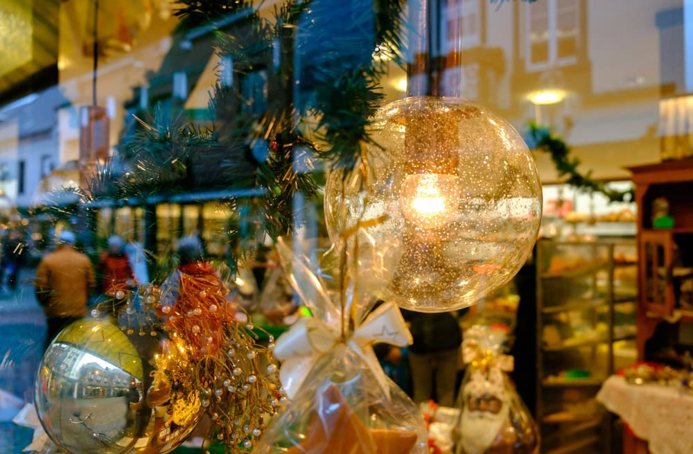 Close-up of Christmas ornaments and decorations hanging in a shop window, with reflections of a street and people outside.