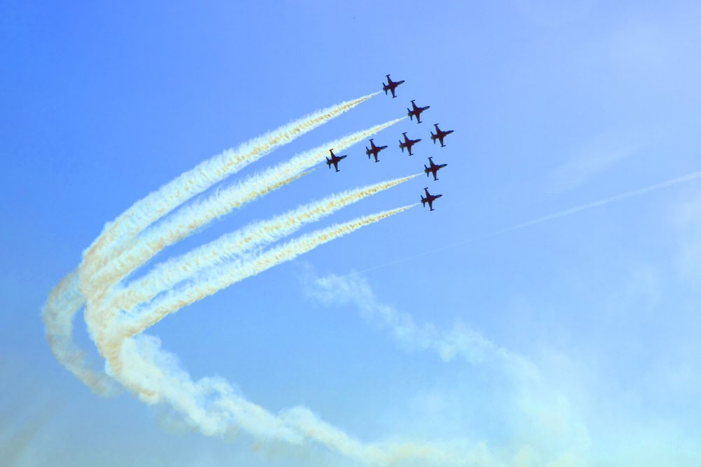 Seven jet planes fly in a tight formation, leaving curved white smoke trails against a clear blue sky.