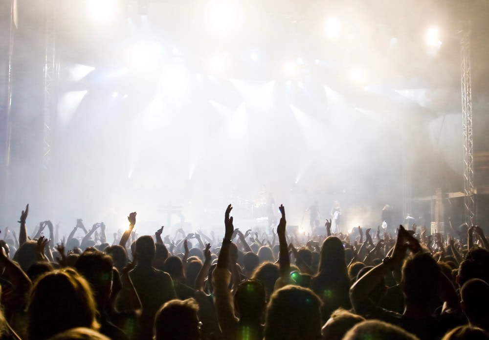 Audience members with raised hands watch a live concert on a brightly lit stage, surrounded by fog and stage lights.