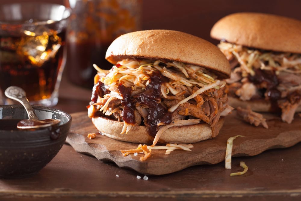 Pulled pork sandwiches topped with coleslaw and barbecue sauce on a wooden board, with a bowl of sauce and a glass of soda in the background.