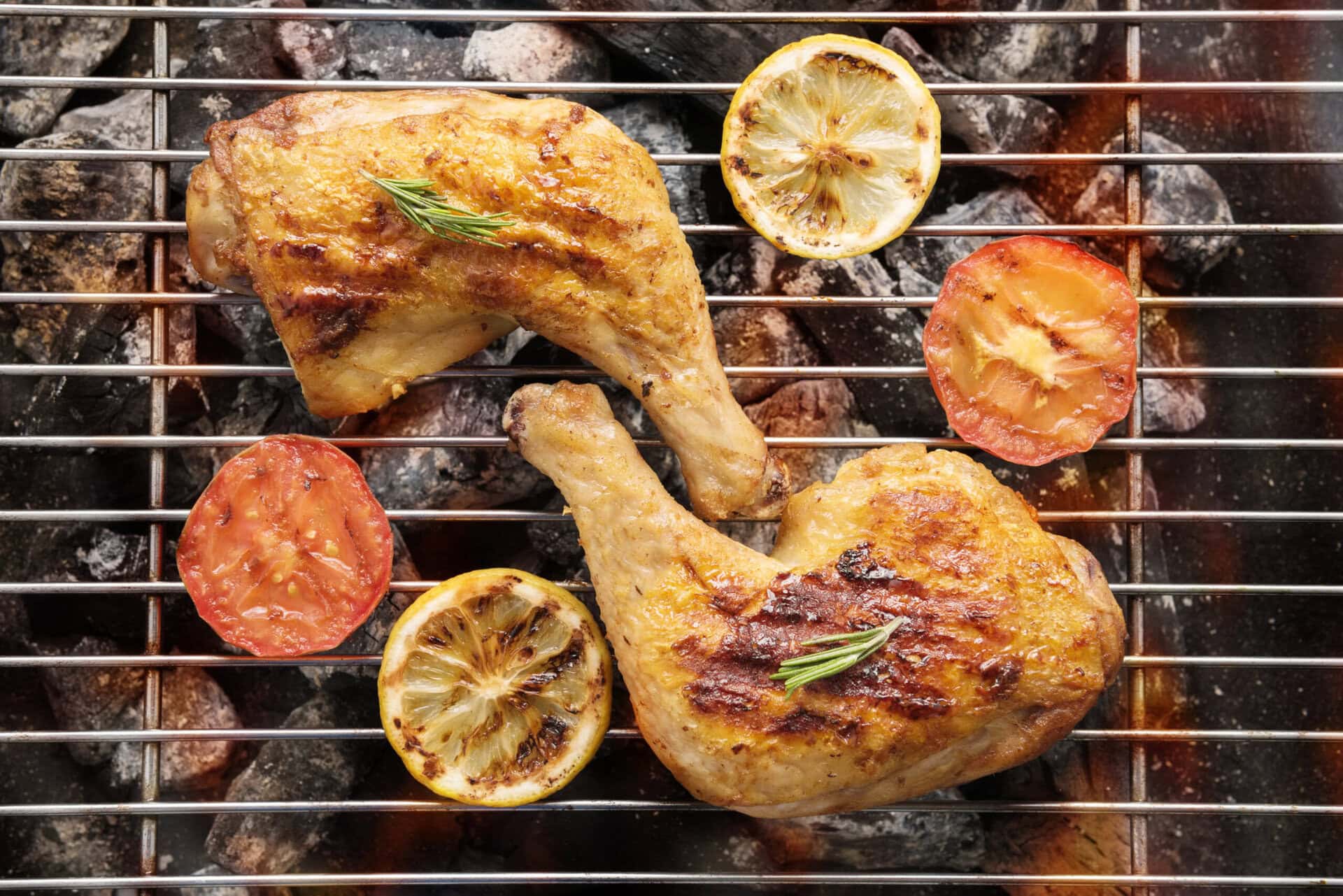 Two grilled chicken legs with rosemary, grilled lemon slices, and tomato halves on a barbecue grill over hot coals.