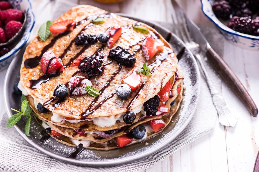 A stack of pancakes topped with mixed berries, mint leaves, powdered sugar, and chocolate drizzle, served on a round plate.