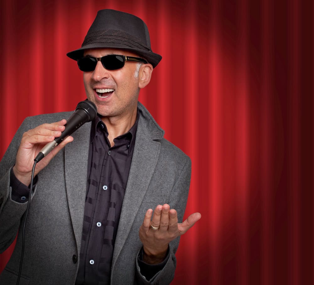 Man in a gray jacket, black shirt, hat, and sunglasses holds a microphone and gestures with his hand, standing in front of a red curtain.