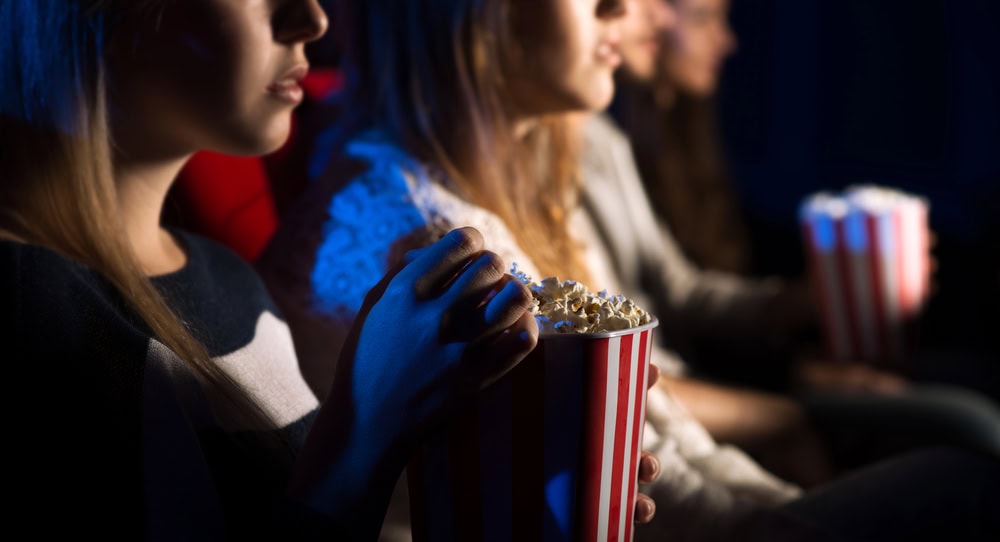 People sitting in a dark movie theater, holding striped popcorn buckets and watching a film.