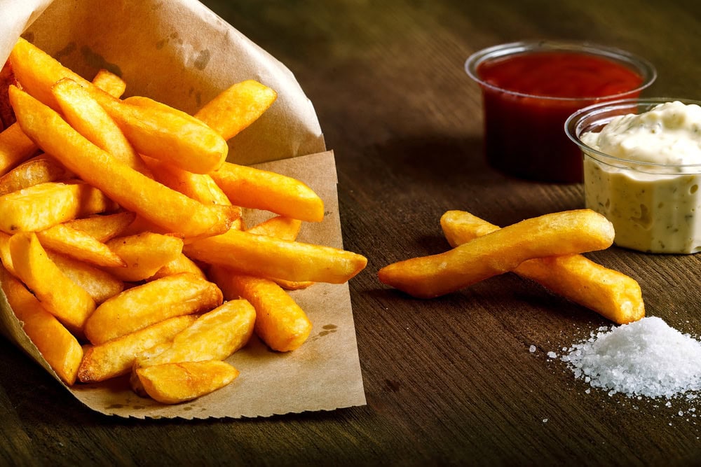 Golden French fries in a paper bag on a wooden surface, with two dipping sauces—one red and one white—and a small pile of salt beside them.