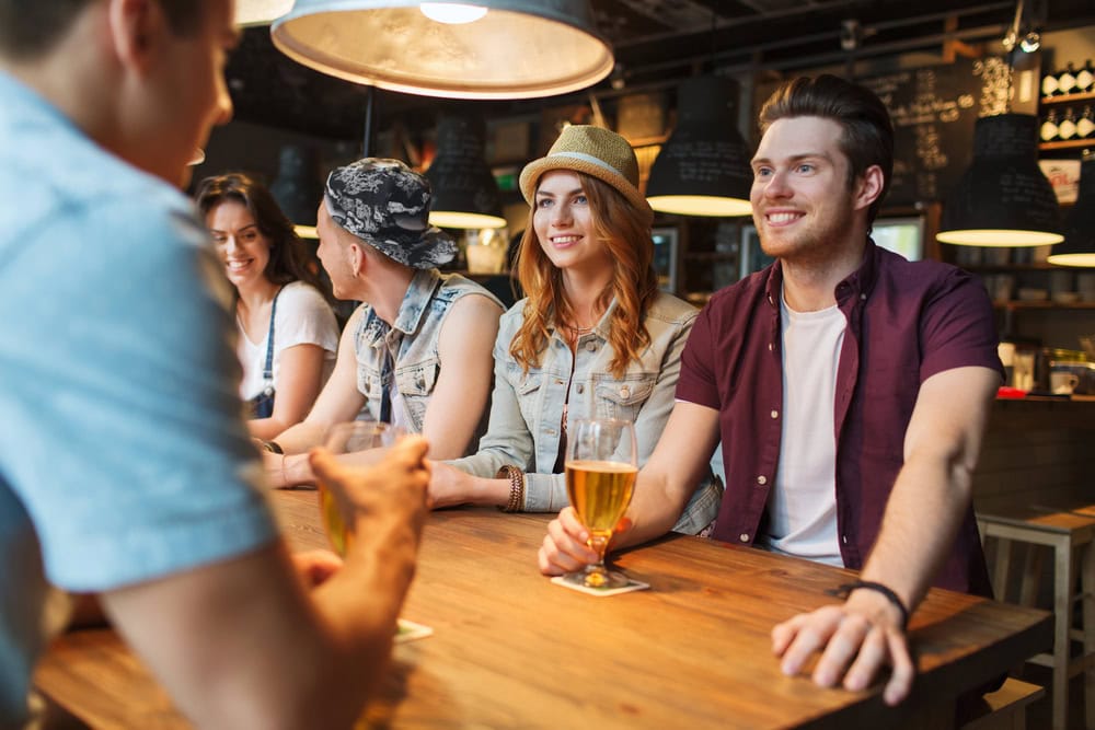 Group of young adults sitting at a wooden table in a bar, smiling and having drinks in a modern, dimly lit setting.