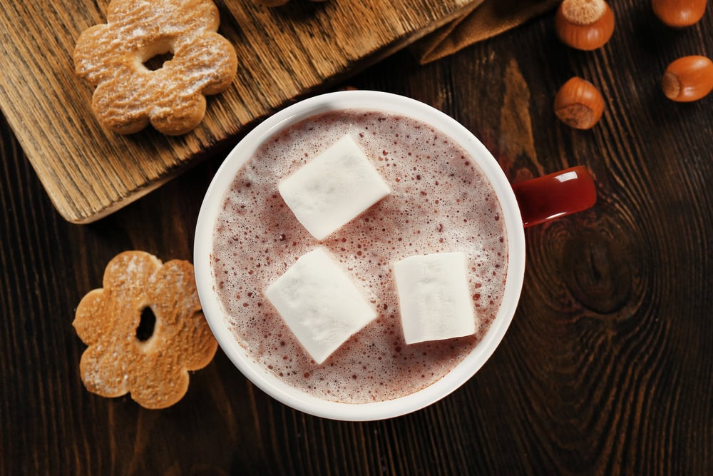A mug of hot chocolate with three marshmallows on top, surrounded by cookies and hazelnuts on a dark wooden surface.