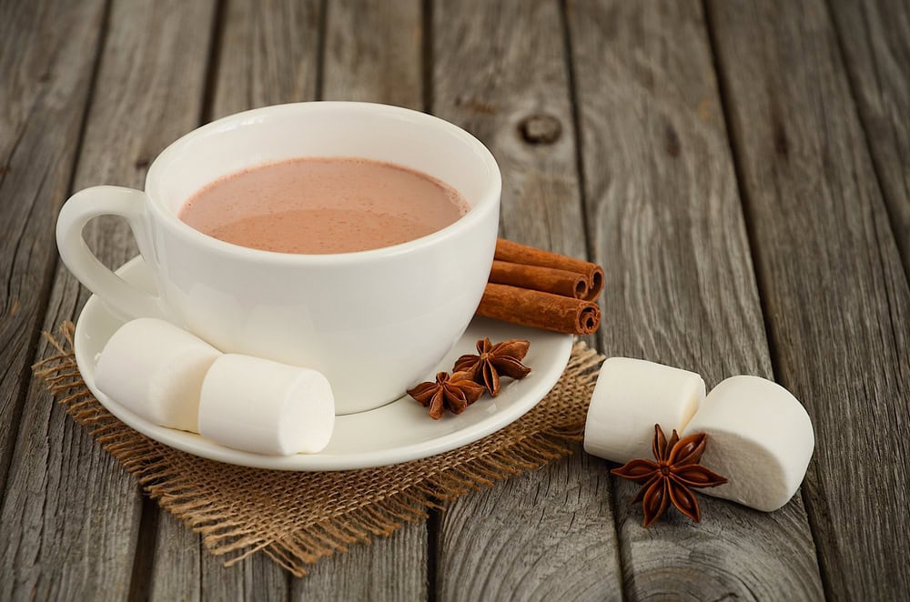 A white cup of hot chocolate on a saucer with marshmallows, cinnamon sticks, and star anise on a wooden table.