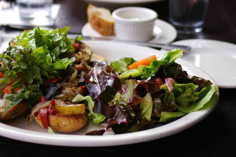 A white plate with a mixed green salad and roasted vegetables on toasted bread, with a slice of bread and a ramekin in the background.