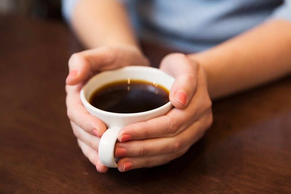 A person holding a white cup filled with black coffee, resting it on a wooden table.