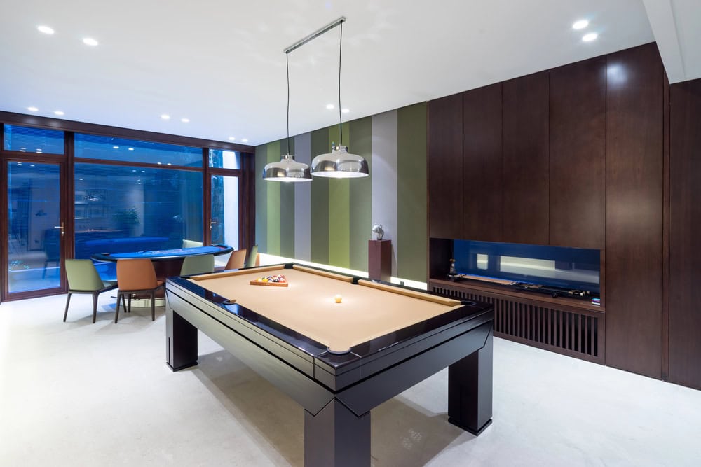 Modern game room with a billiards table, overhead lights, striped accent wall, built-in fireplace, and a seating area near large windows.