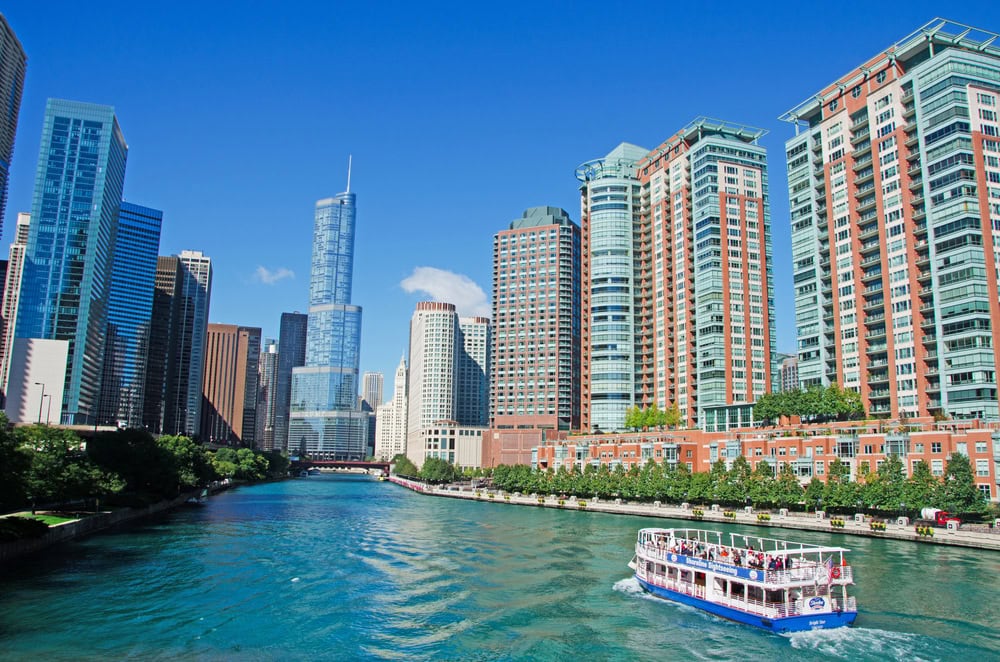 A sightseeing boat travels along a river surrounded by tall modern buildings under a clear blue sky.