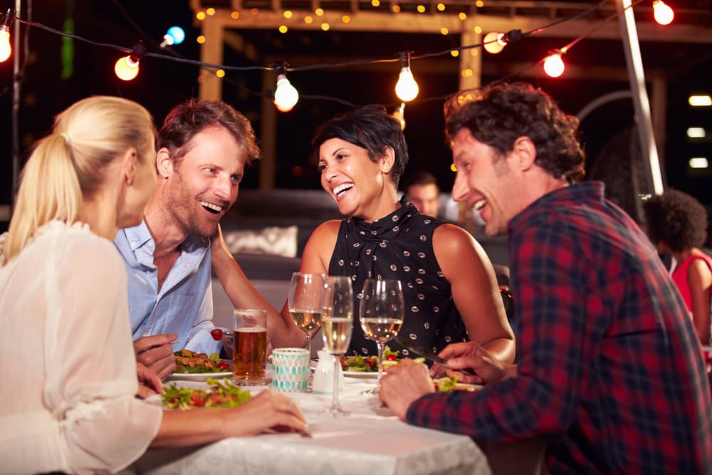 Four adults sit at an outdoor restaurant table eating dinner, drinking wine and beer, and talking together under string lights at night.