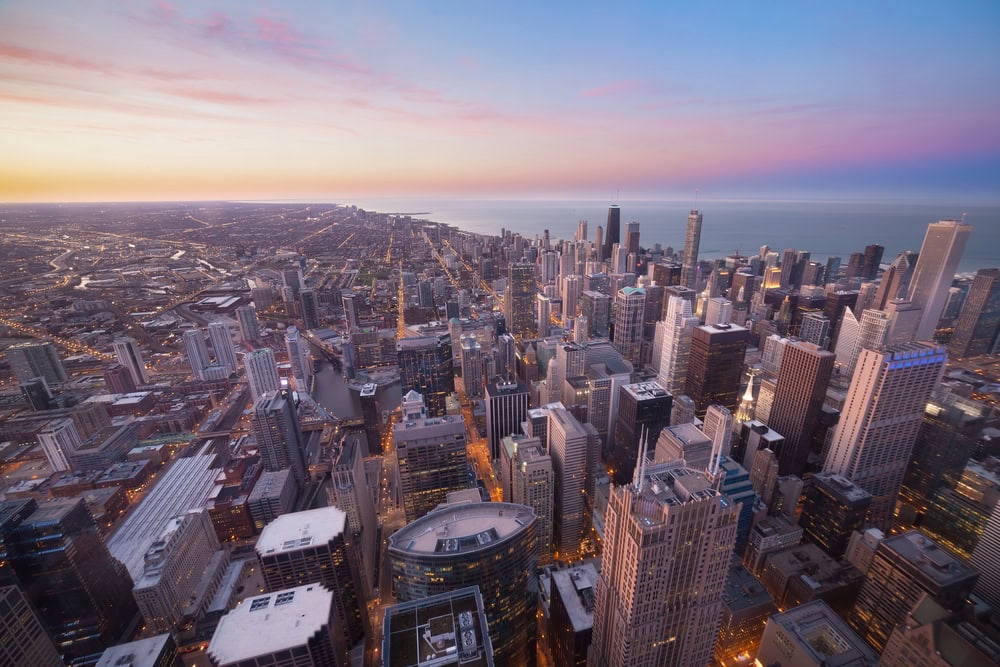 Aerial view of a city skyline with tall skyscrapers at sunset, with Lake Michigan and a colorful sky in the background.