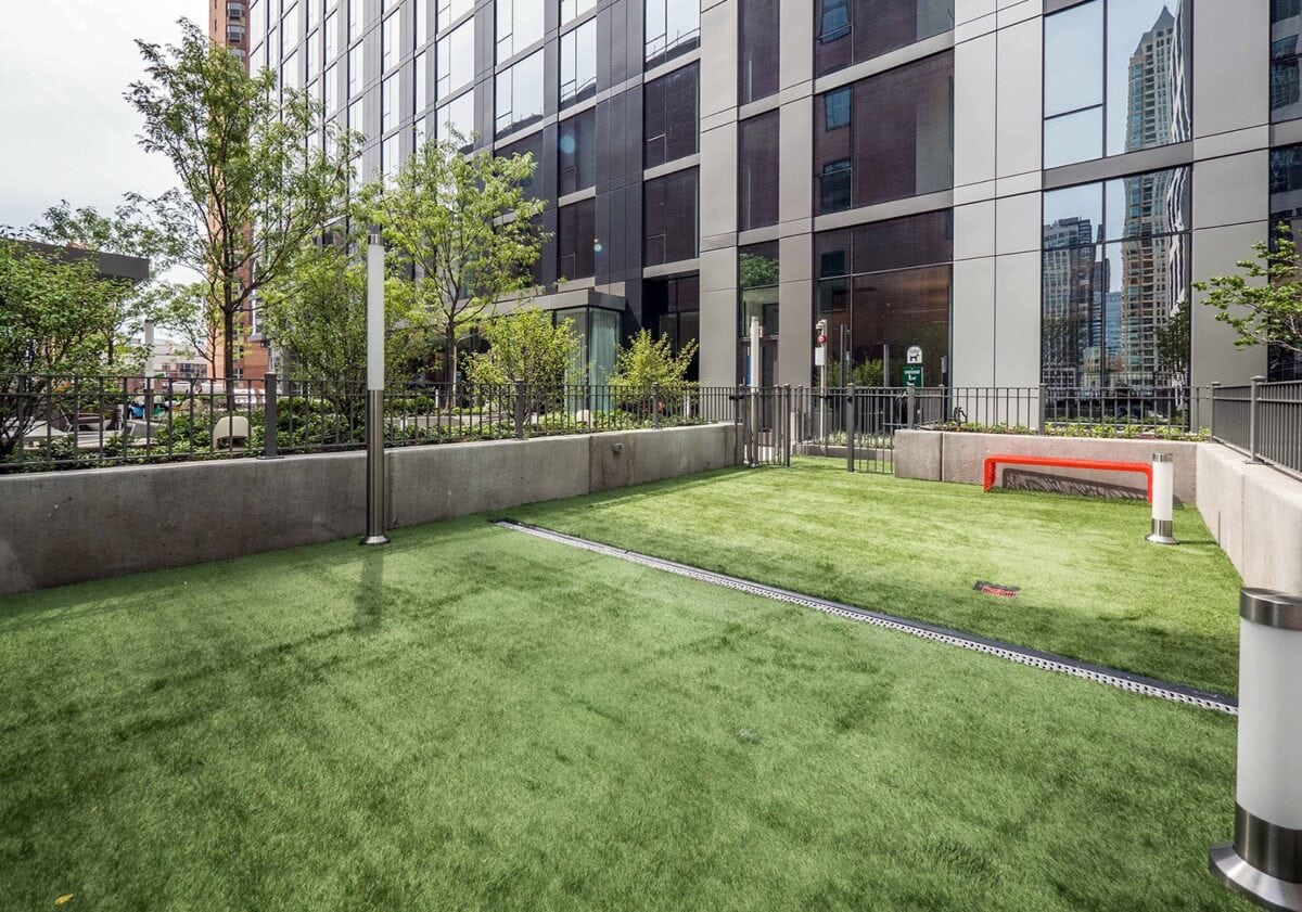 A fenced outdoor area with artificial grass, modern lighting, trees, and a red bench beside a tall building in an urban setting.