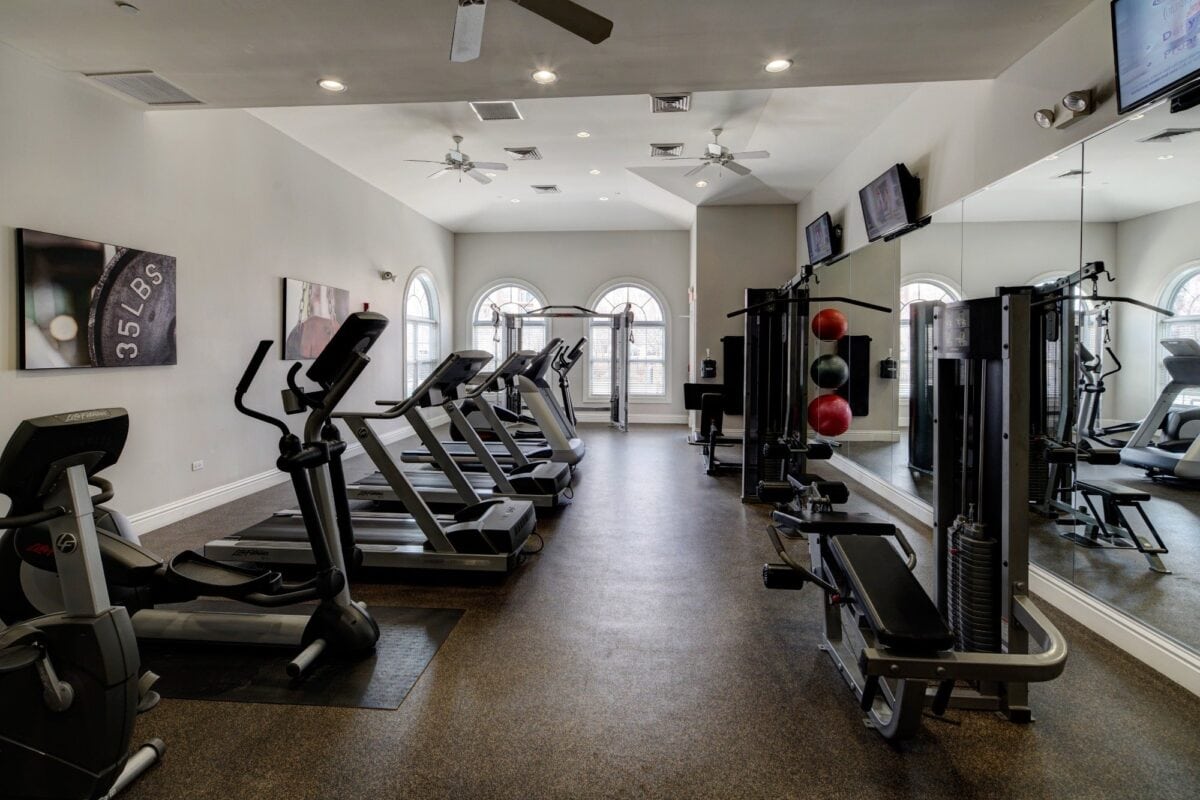 Modern gym with treadmills, ellipticals, weight machines, and large mirrors. Two arched windows, ceiling fans, and wall-mounted TVs are visible.