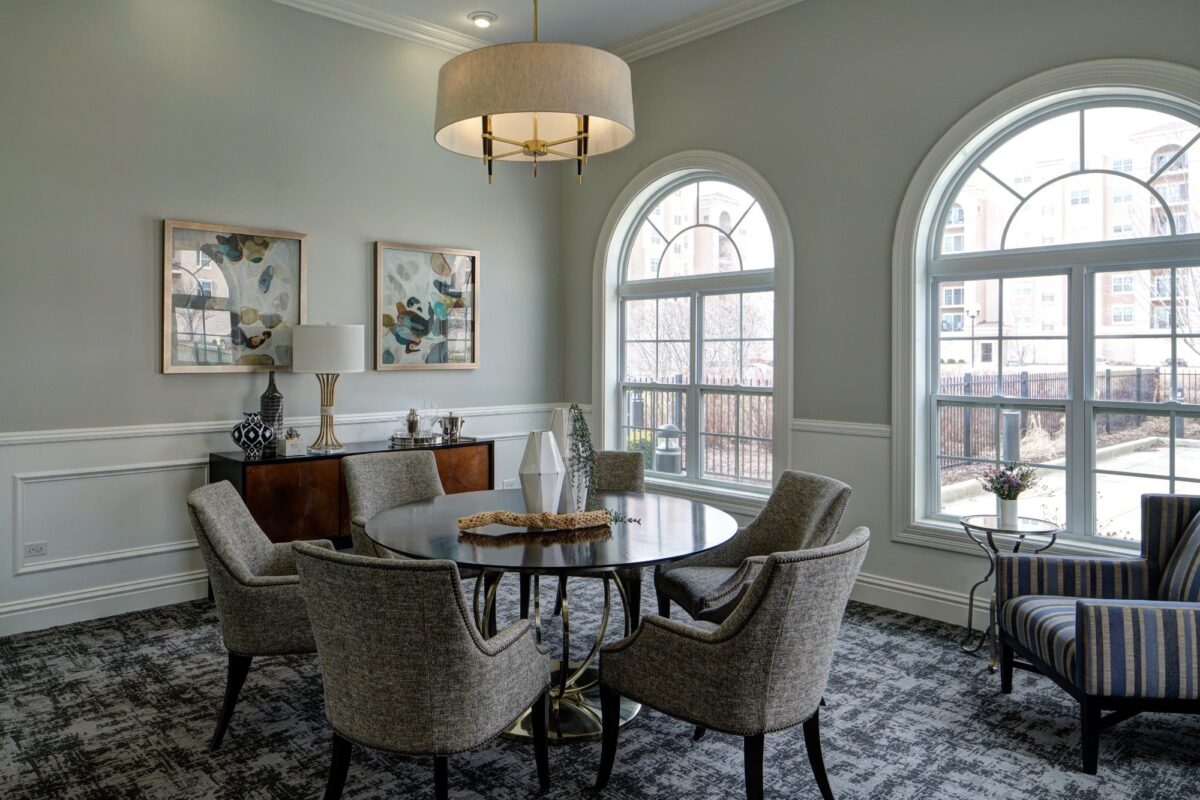 A modern dining room with a round table, six upholstered chairs, two large arched windows, a sideboard, and abstract artwork on the wall.