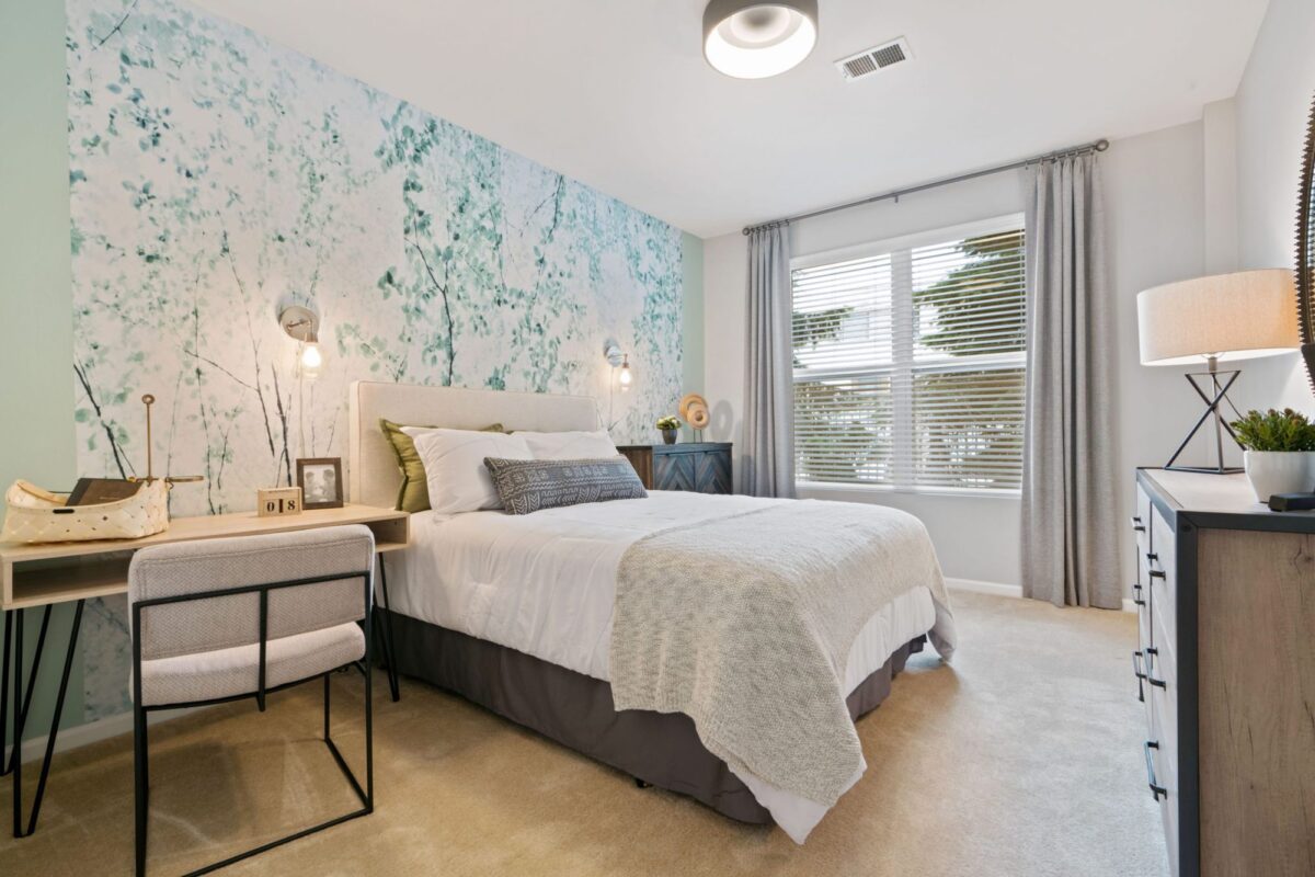 Modern bedroom with a bed, desk, chair, and dresser. Blue and white abstract accent wall, large window with gray curtains, beige carpet, and contemporary lighting.