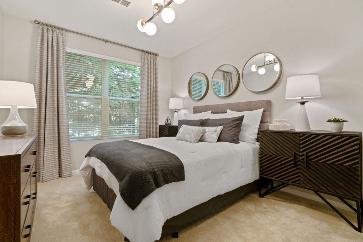 Modern bedroom with a large window, bed with gray and white bedding, two nightstands with lamps, and three round mirrors above the headboard.