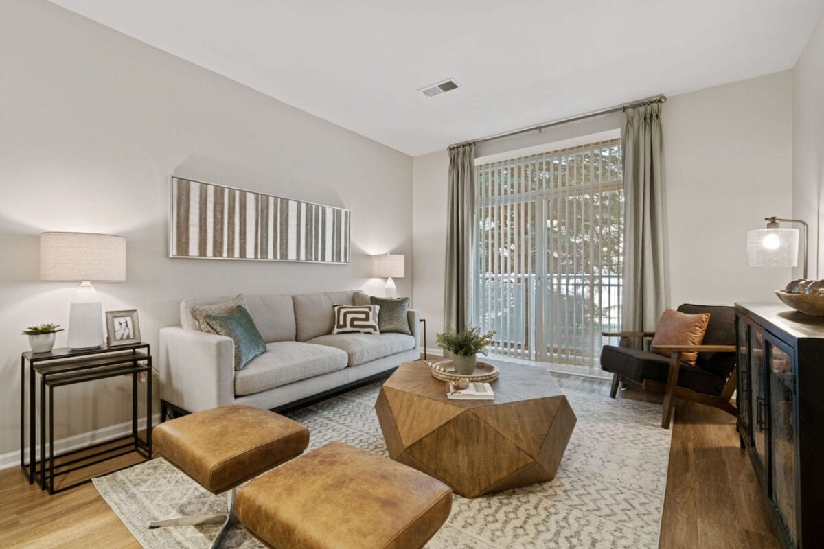 Modern living room with a beige sofa, geometric wooden coffee table, two brown ottomans, accent chair, side tables, lamps, and large window with blinds and curtains.