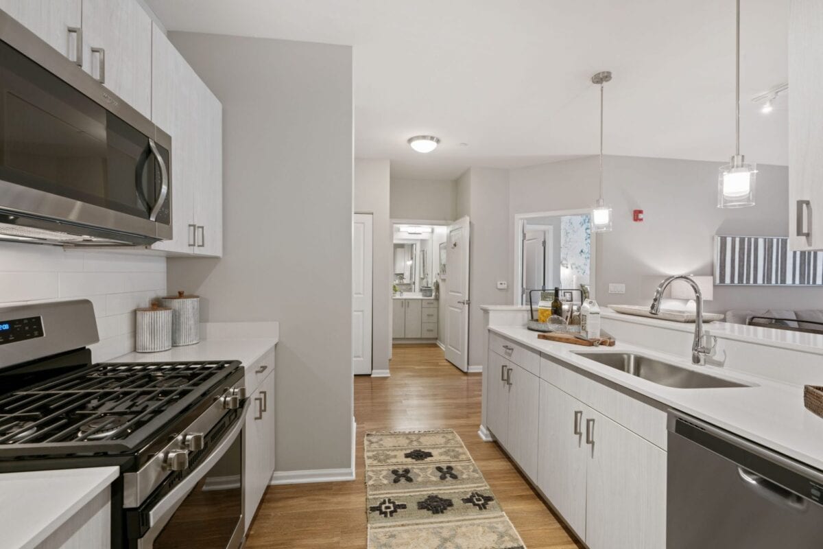 Modern kitchen with stainless steel appliances, white cabinets, a gas stove, double sink, pendant lighting, and a patterned rug on wood flooring. Bathroom visible in the background.