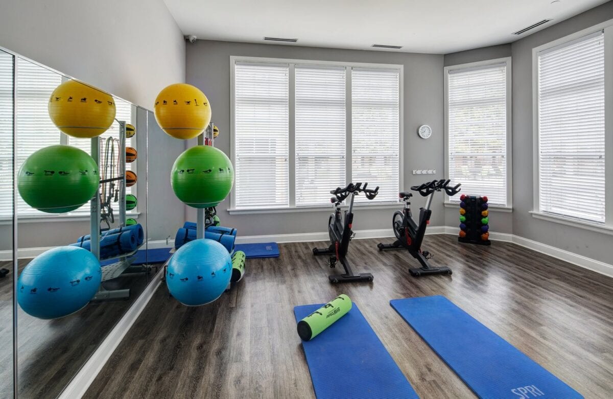 A bright exercise room with two stationary bikes, blue mats, resistance balls, a foam roller, and a rack of weights on a wood floor.
