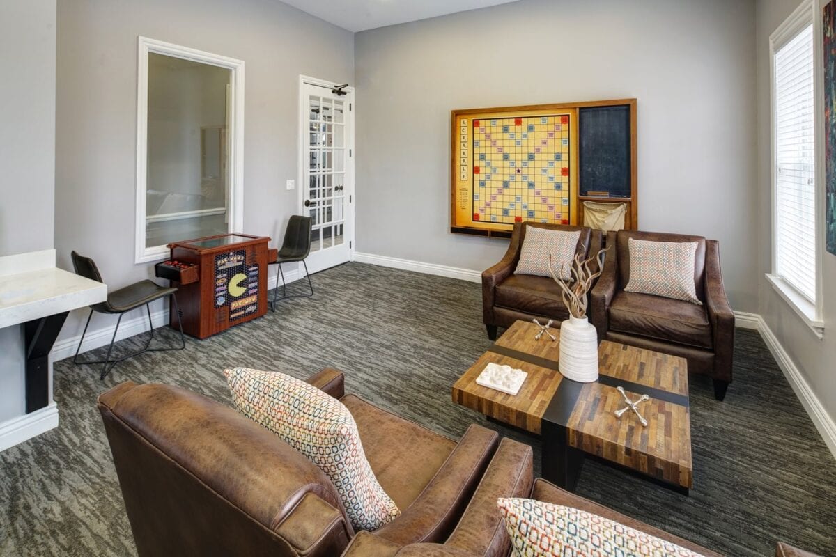 A lounge with two brown leather chairs, a brown sofa, two wooden coffee tables, a large Scrabble board on the wall, and a small desk with chairs near the corner.