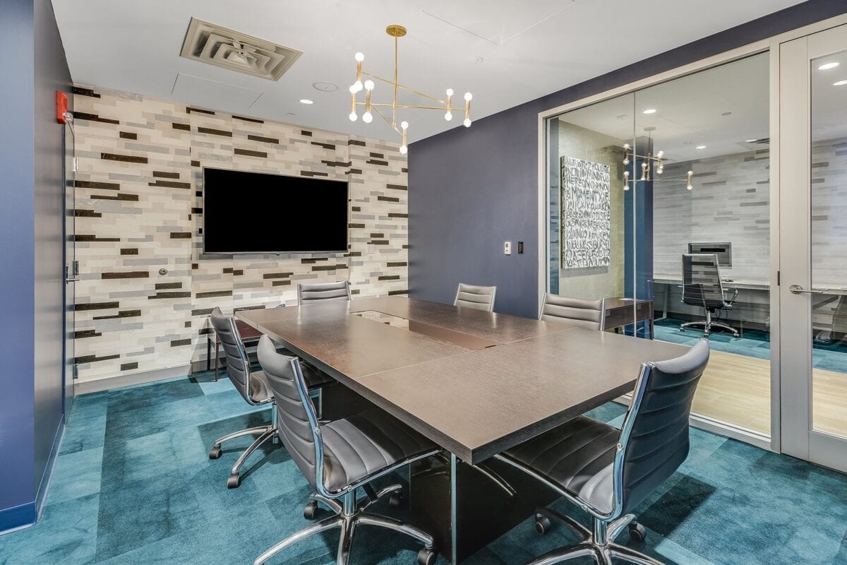 Modern conference room with a rectangular table, six chairs, a wall-mounted TV, and glass partition separating an adjacent office space.