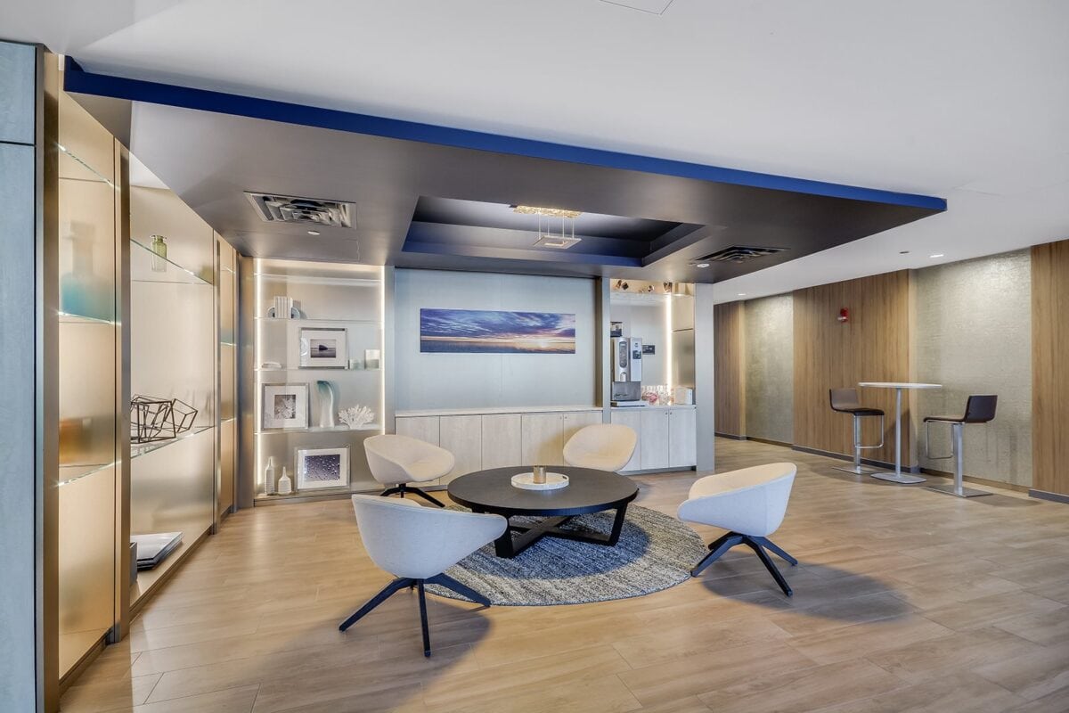 Modern lounge area with four white chairs around a black round table, wall shelves with decor, artwork, and light wood flooring.