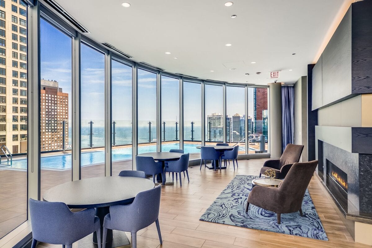 Modern lounge with floor-to-ceiling windows, blue chairs, round tables, and a fireplace, overlooking a pool and cityscape with tall buildings and a view of the water.
