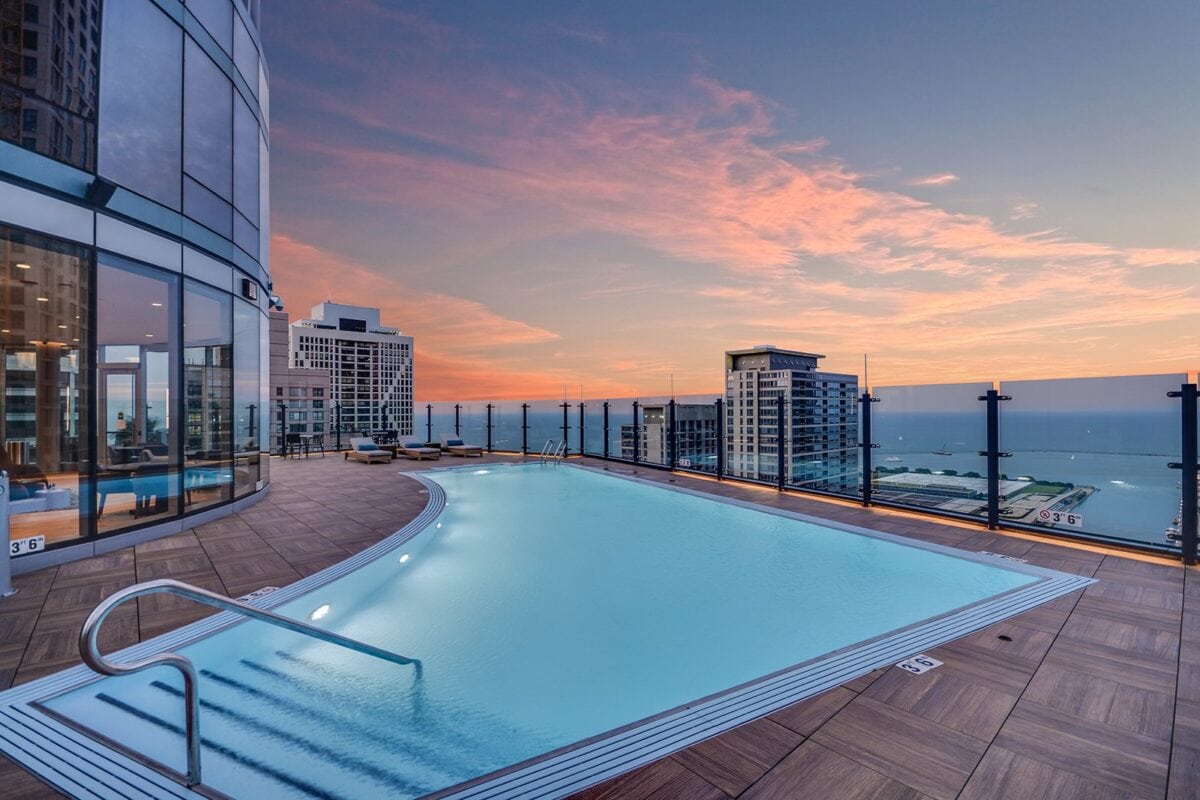 Rooftop infinity pool with glass railing overlooking city buildings and water at sunset, reflecting the pink and blue sky.