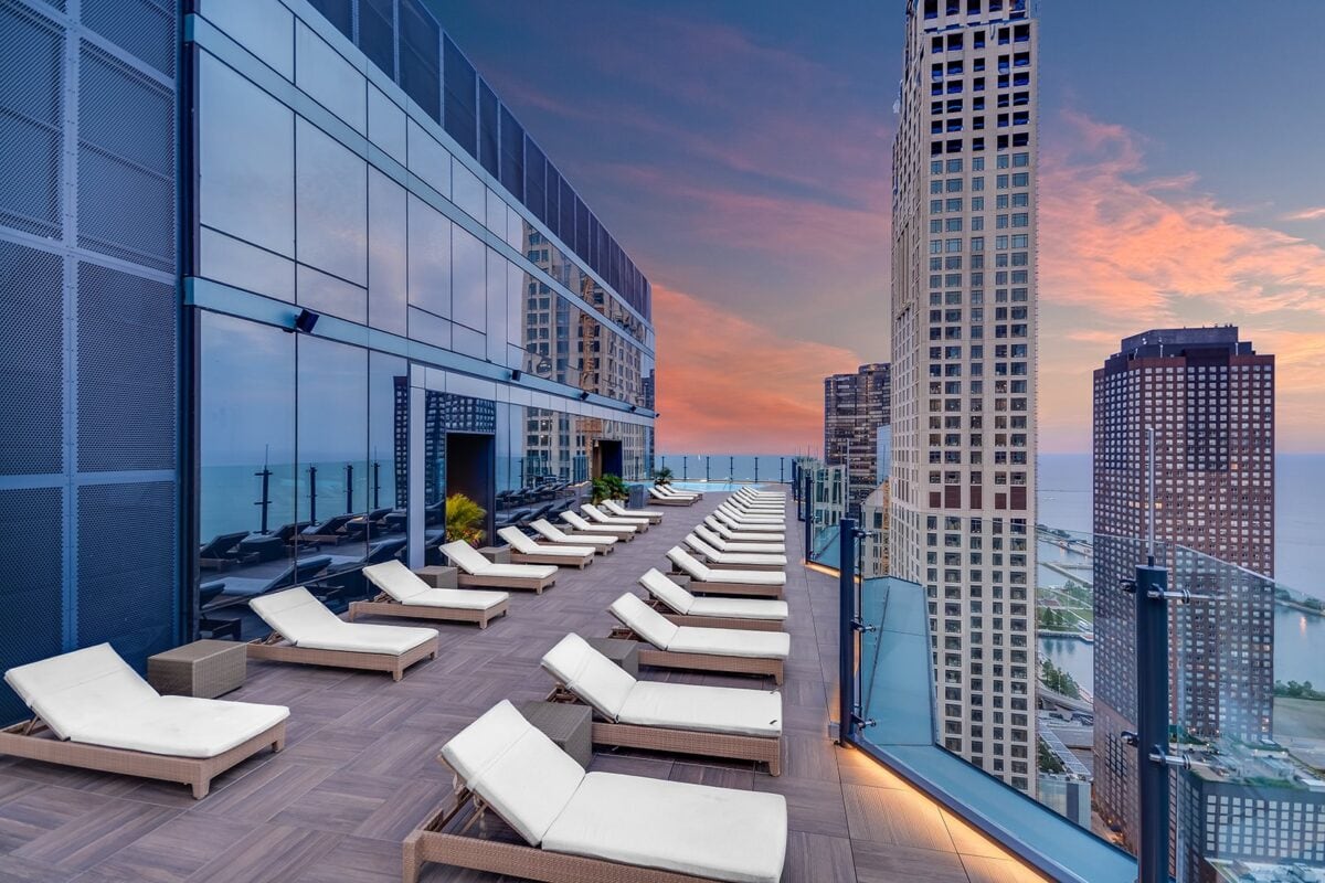 Rooftop terrace with rows of white lounge chairs, glass railing, and city skyscrapers at sunset overlooking a body of water.