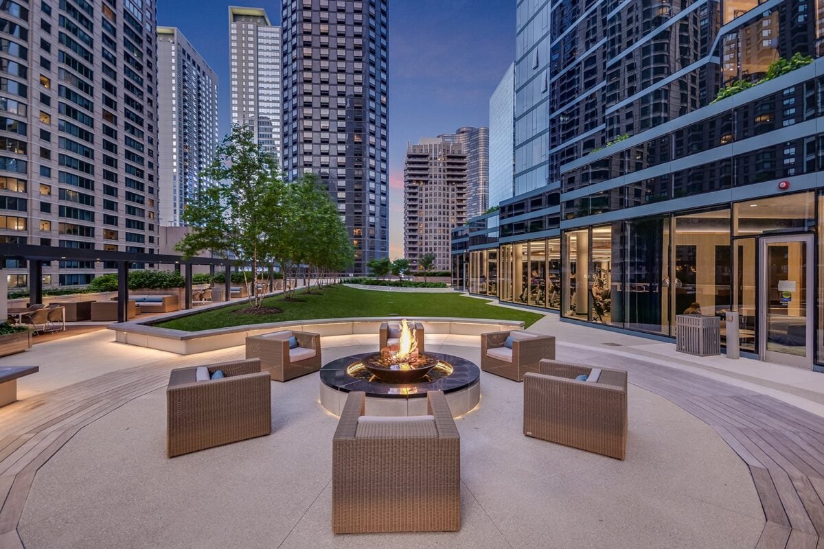 Rooftop terrace in a city with wicker chairs arranged around a fire pit, surrounded by tall modern buildings and landscaped greenery.