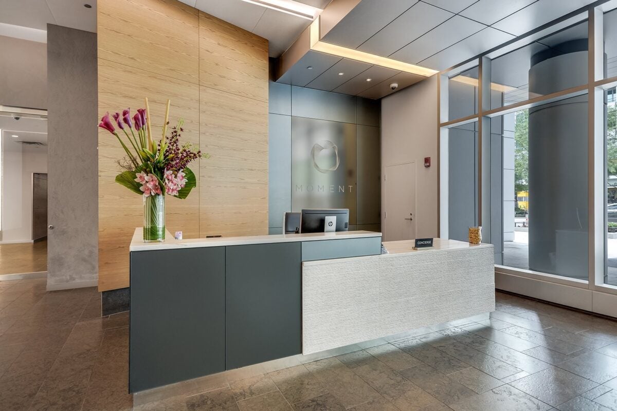 Modern office reception area with a front desk, two monitors, a vase of flowers, and large windows letting in natural light. A logo and the word "Moment" are on the wall behind the desk.