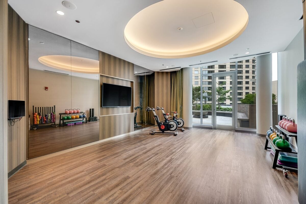 Modern fitness room with wooden flooring, exercise bikes, free weights, mirrored wall, large TV, and floor-to-ceiling windows overlooking an outdoor area.