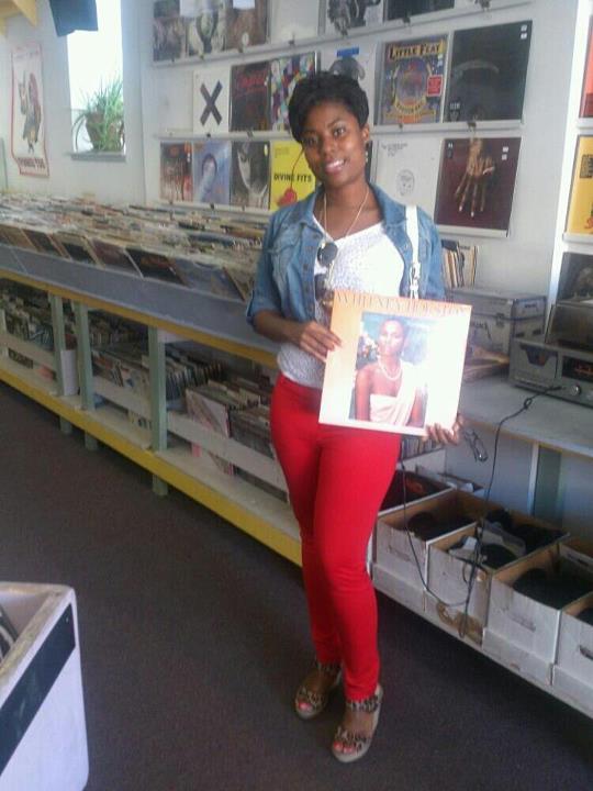 A woman in red pants stands in a record store, smiling and holding a vinyl record with a woman's portrait on the cover.