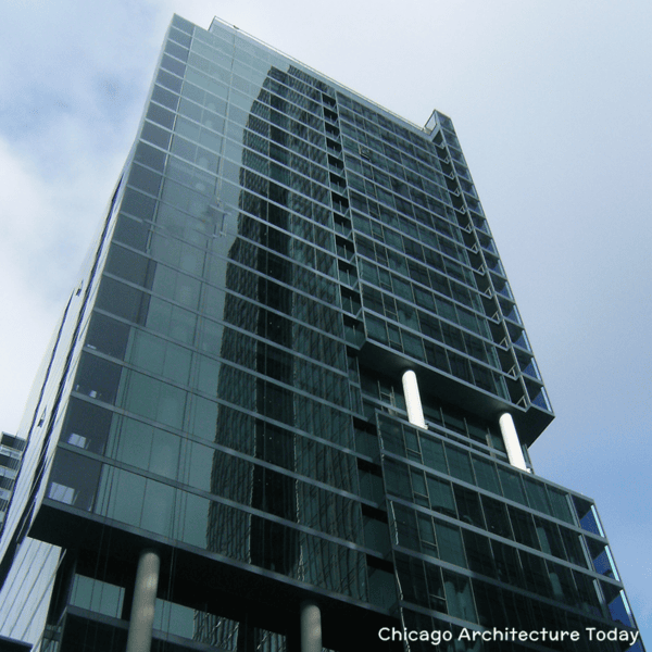 A modern glass high-rise building with reflective windows, viewed from below against a partly cloudy sky. Text in the corner reads 