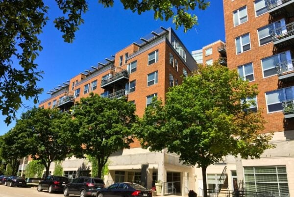 Red brick apartment building with balconies and large windows, lined by parked cars and leafy green trees under a clear blue sky.