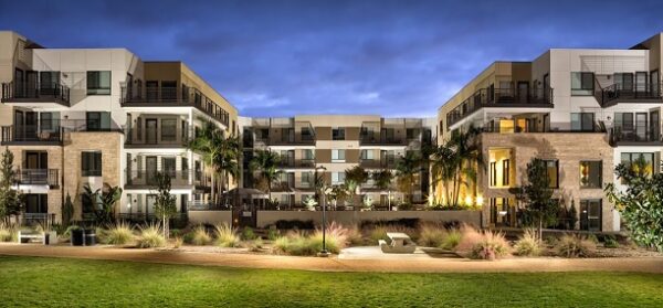 Modern apartment complex at dusk with illuminated windows, balconies, palm trees, and landscaped grounds featuring pathways and benches in the foreground.