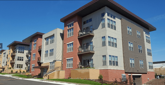 A row of modern, multi-story apartment buildings with balconies, large windows, and a mix of brick and siding exteriors under a clear blue sky.