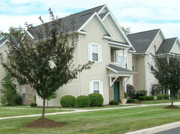 Two beige two-story suburban houses with pitched roofs, green doors, and small front lawns, separated by landscaped shrubbery and sidewalks.