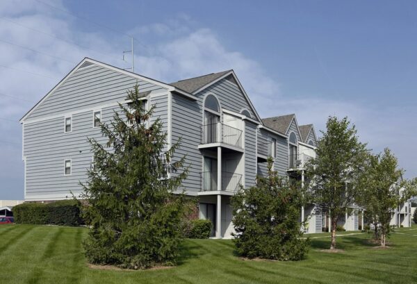 A row of modern, gray three-story apartment buildings with balconies, surrounded by green grass, trees, and shrubs under a clear sky.